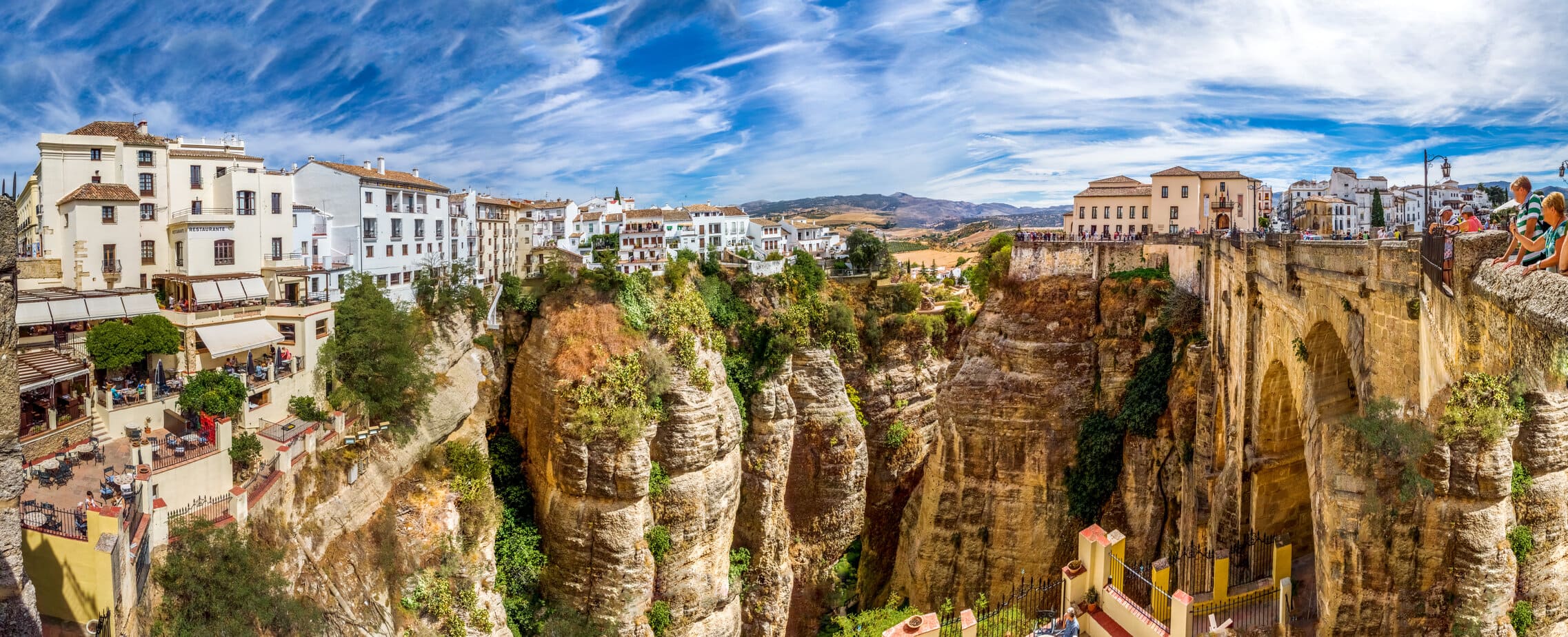 panorama of the Ronda in Andalucia Spain on a very sunny day with blue sky above. the town is on the top of a sheer rock cliff with a stone bridge on the right of the image. best day trips from Malaga. 