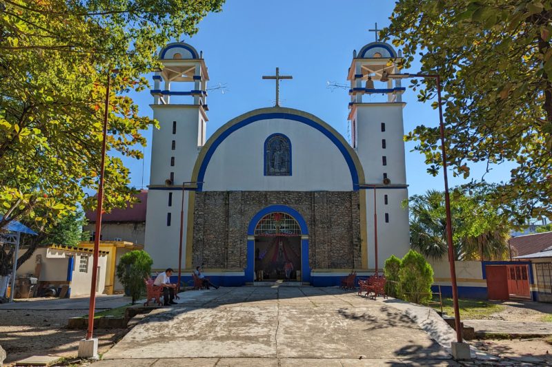 front of a small mexican church with a domed roof with a cross on top and two small towers either side. most of the facade is white with bright blue trim, the lower half of the centre of the facade is exposed grey stone with a blue arched doorway in the middle.