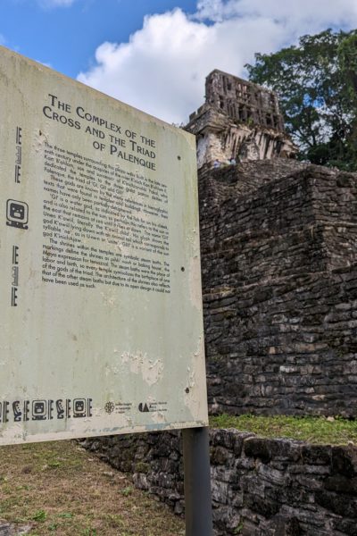 white sign with the heading "The Complex of the Cross and the Triad of Palenque". Behind the sign is a large grey stone pyramid out of focus.