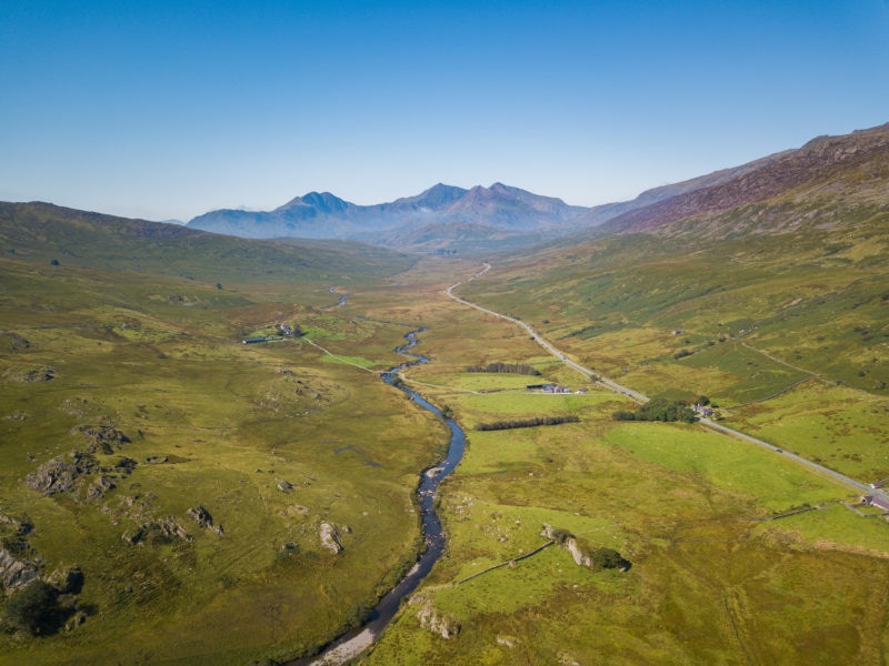 aerial shot of a river and a road in a wide grassy valley with a sloping hill on either side and three mountain peaks in the distance