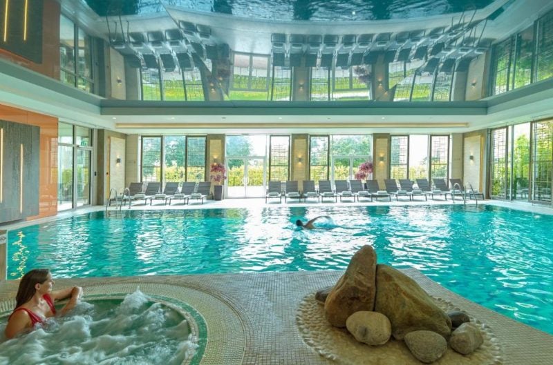 a lady in a red swimsuit sitting in a hot tub next to a large blue indoor swimming pool with large windows on te far side looking out onto a green lawn. Sopwell House in St Albans England