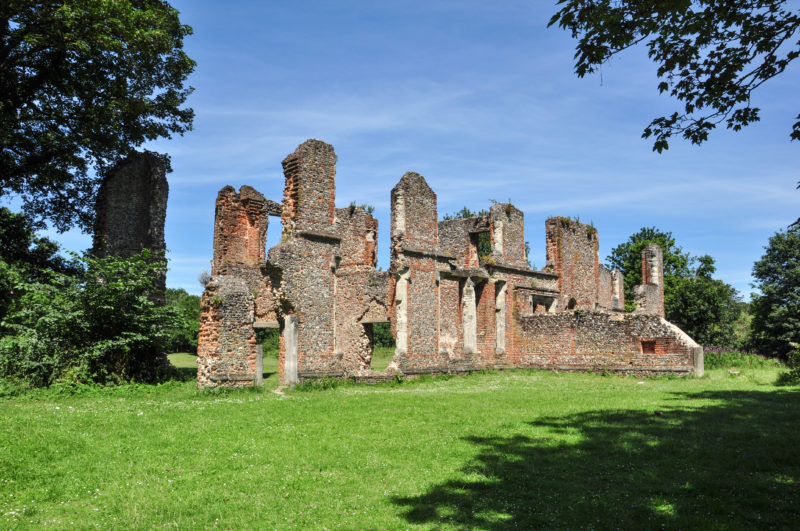 ruins of an old nunnery building with ruined red brick walls and empty windows standing in a grassy field with a few trees visible on either side on a sunny day with blue sky. things to do in st albans england. 