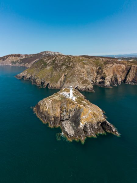 aerial view of a small rocky islet in the blue sea off the coast of anglesey in north wales with a white lighthouse tower on the islet and the island of anglesey in the background behind it. 