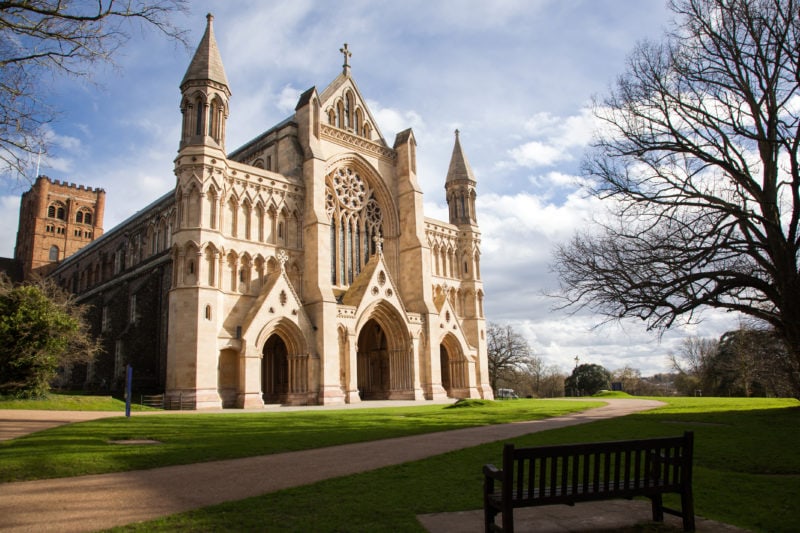 St Albans Cathedral on sunny day in winter with a grassy lawn and bare tree in front. the cathedral is cream coloured and has three arches at the bottom of the carved front facade. 