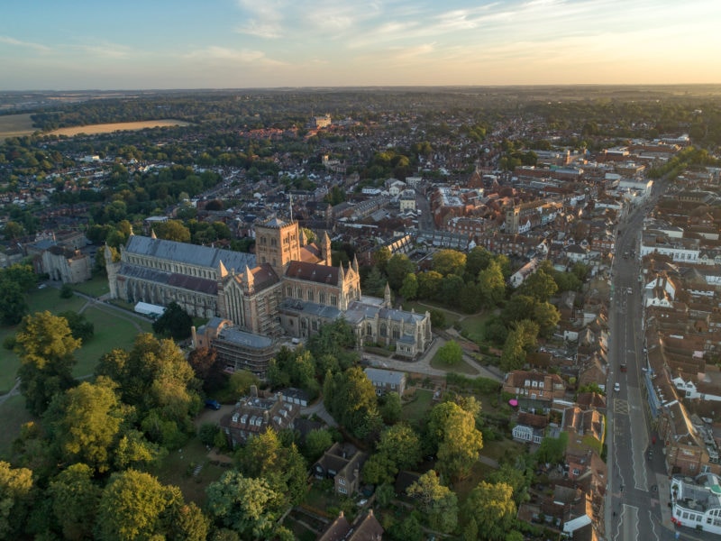 aerial shot of St Albans in England with the cathedral at the front and a large wooded area in front of that and the town behind, taken at sunset with a golden glow in the sky above. things to do in St Albans England. 
