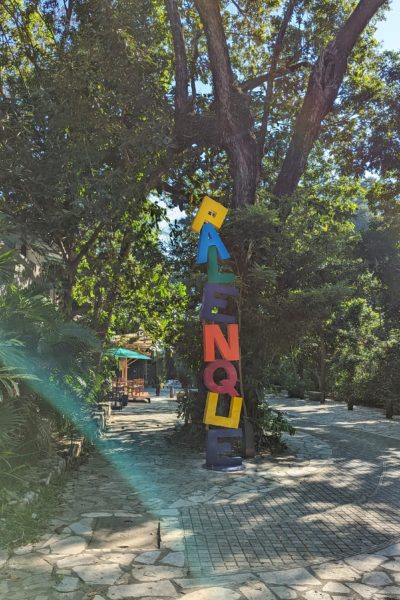 flagstone street with dense leafy trees on either side and a vertical sign with different coloured letters spelling out the word "Palenque" 