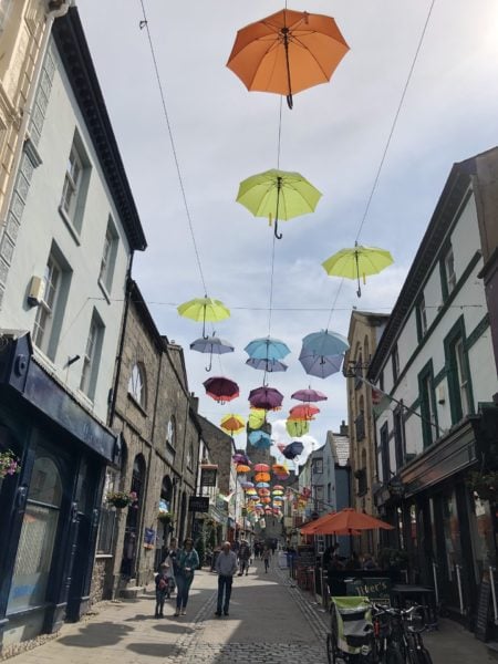 looking down a busy high street in a town with strings of different coloured umbrellas suspended overhead against a cloudy sky - things to do in Caernarfon Wales