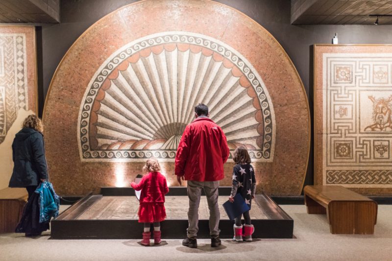 man in red jacket with two young girls on either side of him seen from behind looking at a large circular roman mosaic inside a museum.