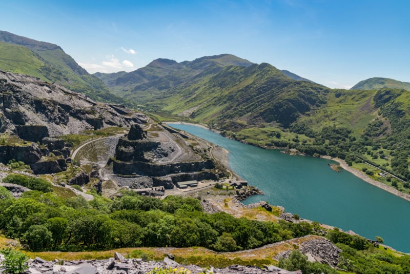 Looking down from a tall hill at a bright blue lake withe the remains of a grey slate quarry in the cliffs on the left on a bright sunny day in Llanberis Wales with blue sky overhead