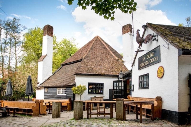 exterior of a pub with white washed walls and a grey pointed roof with two very tall white painted chimneys on either side. the sign reads 'ye olde fighting cocks'. it is a sunny day with blue sky overhead and there are wooden tables outside the pub. 