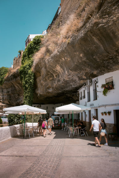 a white washed resturant buidling built into the base of a rock cliff with a patio outside with several tables and white umbrellas on a very sunny day with blue sky above. Best day trips from Malaga. 