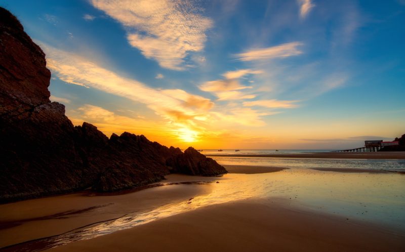 an empty flat beach at sunrise with the sun coming up over the sea and very shallow still water on the sand of the beach. there are some large rocks silhouetted on the left and a glimpse of the town of tenby in the distance on the right. best wales road trips. 