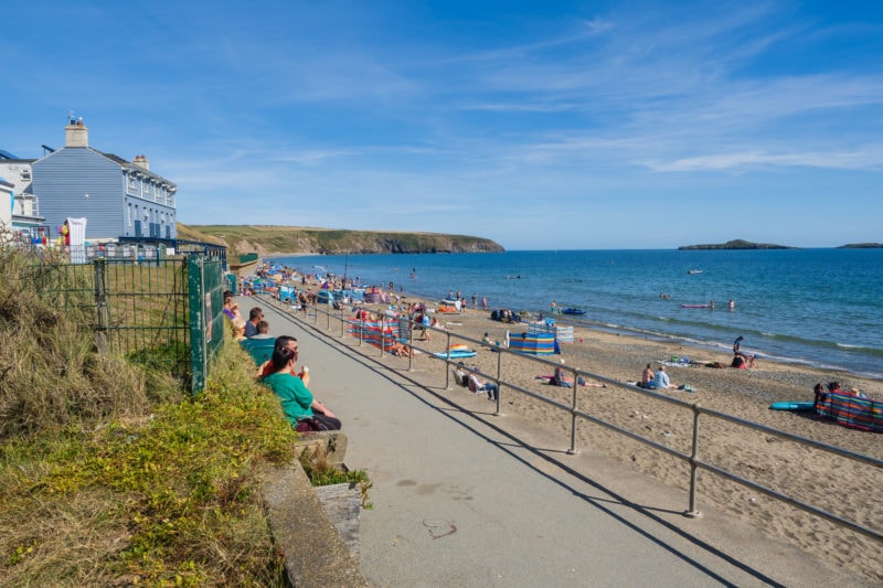 grey paved walking path with metal railings nexct to a long sandy beach wit bright blue sea. there are a few people sitting on benches on the left side of th epath looking out to see, the closest woman is wearing a green t shirt and looking away from the camera. in the distance to the left are a few buildings of the small village of Aberdaron and in the farther distance is a low green headland