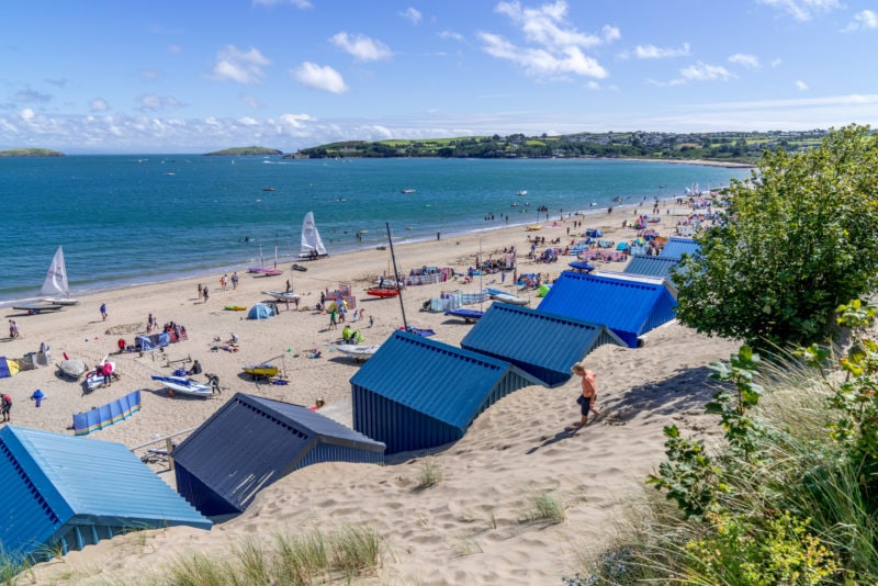 long white sandy beach viewed from behind a row of small beach huts with blue roofs. the beach is crowded with people lying on towels and the sea in front of them is bright blue on a sunny day with blue sky overhead and a couple of small fluffy white clouds. best beaches on the llyn peninsula