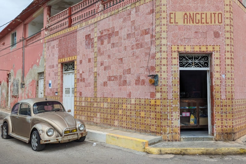 an old golden coloured VW beatle car parked in the street outside an old one storey building covered in pink and yellow tiles with the words El Angelito written in tiles above a doorway on the corner of the building.