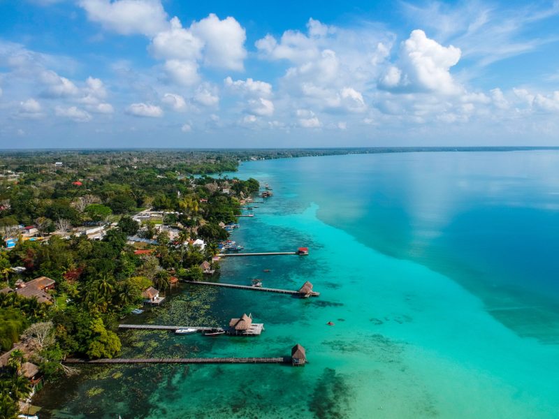 aerial photo of a huge lagoon with striking turquoise water and the shore covered in green tropical forest with several long wooden jetties reaching out into the lake - Bacalar travel guide Mexico
