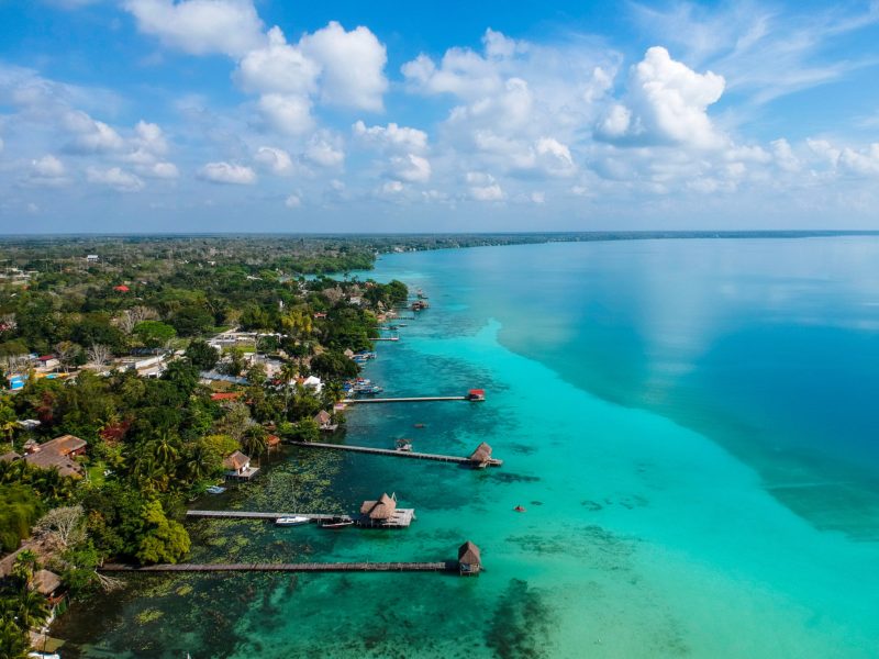 aerial photo of a huge lagoon with striking turquoise water and the shore covered in green tropical forest with several long wooden jetties reaching out into the lake - things to do in bacalar mexico