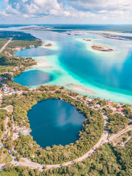 aerial photo of a large landscape with a cyan lake and three circular cenotes with dark blue water - two are under the lake and the closest one is just next to it surrounded by green forest - bacalar lagoon travel guide mexico