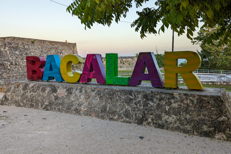 low grey stone wall topped with different coloured letters spelling out the word bacalar with the edge of a grey stone fort in the background near sunset with a washed out pink sky above