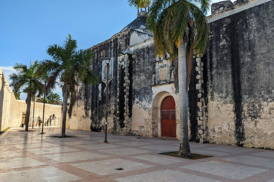 part of the old city walls in campeche with a dark red wooden door set into an archway in the grey stone walls. there is a new plaza in front with creamy coloured tiled floor and a line of palm trees on a very sunny day with blue sky above