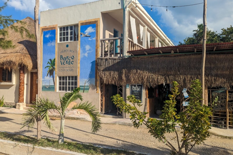 exterior of a whitewashed two storey block shaped building with a single storey bar with grass roof alongside it. the wall of the white building has a large painting of the blue sky with the name Barco Verde Hostel painted on it, and the hostel is next to a sandy road lined with palm trees.