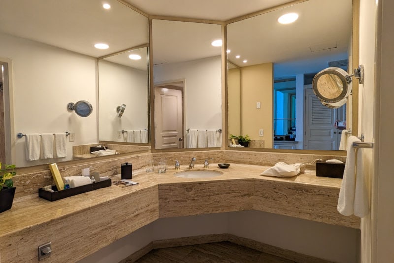 hotel bathroom with a large wraparound marble counter around the corner of the room and a sink in the centre with three large mirrors on the walls above
