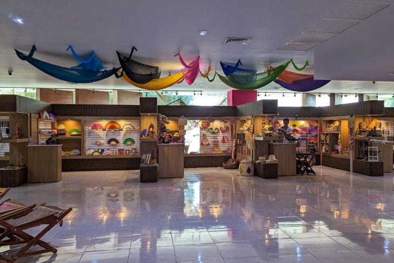 interior of an indoor market place with a shiny white floor and white cieling and a row of wooden stalls all full of different local handicrafts. there are several different colour hammocks hanging from the cieling.