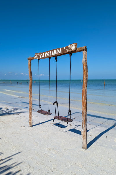 wooden frame with two swings and the word Carolinda painted in white at the top on a white sandy beach with the clear shallow sea behind on a very sunny day with blue sky 