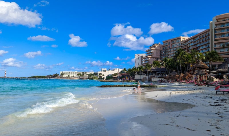 white sandy beach with vivid cyan water and tony waves lapping at the sand. there are sun loungers in the distance and a 12 storey pinkish hotel building visible behind the palm trees to the right.  Grand Fiesta Americana Coral Beach Cancun review. 