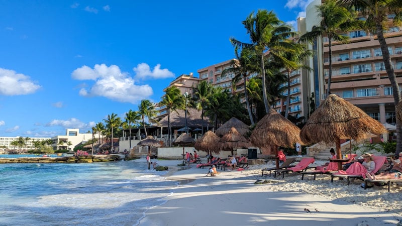 white sandy beach with vivid cyan water and tony waves lapping at the sand. there are sun loungers in the distance and a 12 storey pinkish hotel building visible behind the palm trees to the right. Grand Fiesta Americana Coral Beach Cancun review.