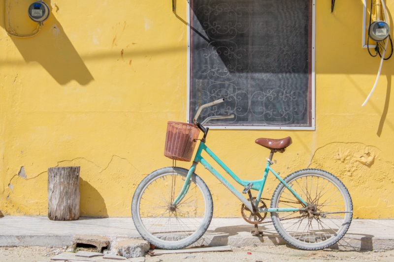 turquoise bike with a wicker basket and brown seat parked at the side of the road next to a bright yellow wall in holbox mexico
