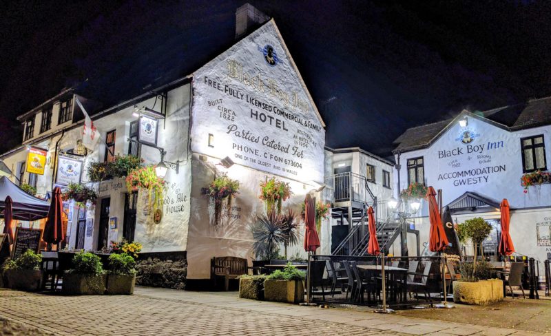 Exterior of the Black Boy Inn in Caernarfon, Wales at night, the inn is painted white and lit up against the dark sky with a few wooden tables on the stone patio area outside