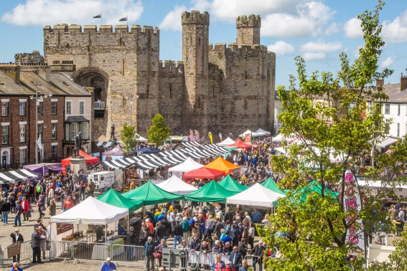 caernafon castle an old stone castle with two turrets with a huge crowd of people and some green and white covered stalls in the square outside it viewed from above during caernarfon food festival