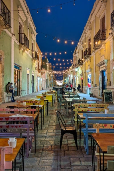 looking along a steet in campeche centro historico at night with colourful buildings on either side and strings of large fairylights overhead against the dark sky. the grey flagstone street is full of painted wooden tables and chairs.
