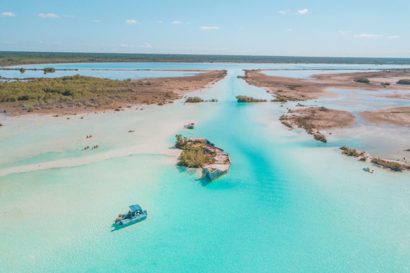 aerial photo of a short channel between two lagoons with vivid cyan water and wild, green land on either side. there is a small boat in the canal and a ruined building on a tiny island in the middle. things to do in bacalar mexico. 