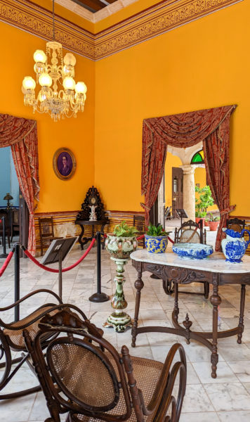 interior of a historic living room in a colonial mansion in Campeche Mexico with wooden rocking chairs and a small table topped with blue and white china. the walls are painted bright yellow and the two doors on either side are surrounded by large red curtains.