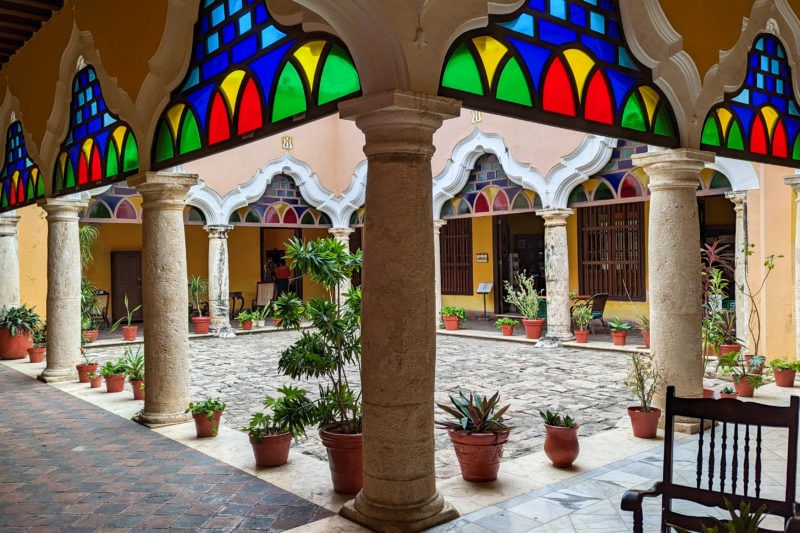 central courtyard of Casa 6 in Campeche with a grey flagstone patio surrounded by yellow columns and arches topped with stained glass windows in green blue and yellow