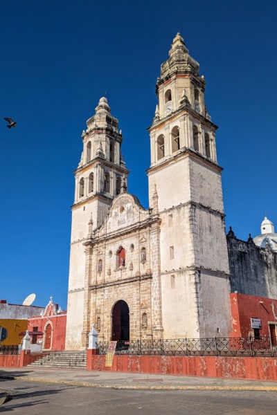campeche cathedral a white stone building with two towers on either side of the front facade with a low red wall along the edge of the street in front with a bright blue sky overhead