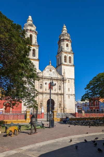 campeche cathedral a white stone building with two towers on either side of the front facade with a curved stone pedestrian path in front and trees on either side with a bright blue sky overhead