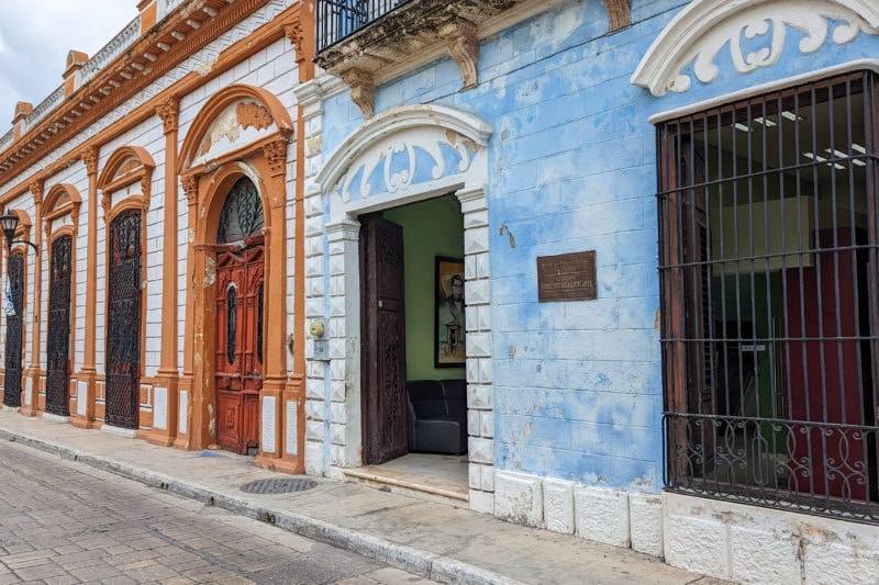 two old colonial style one storey buildings on a street in Campeche Centro Historico, the closest building is painted light blue with cream swirls above the door and window and the further building is painted white and dark orange.