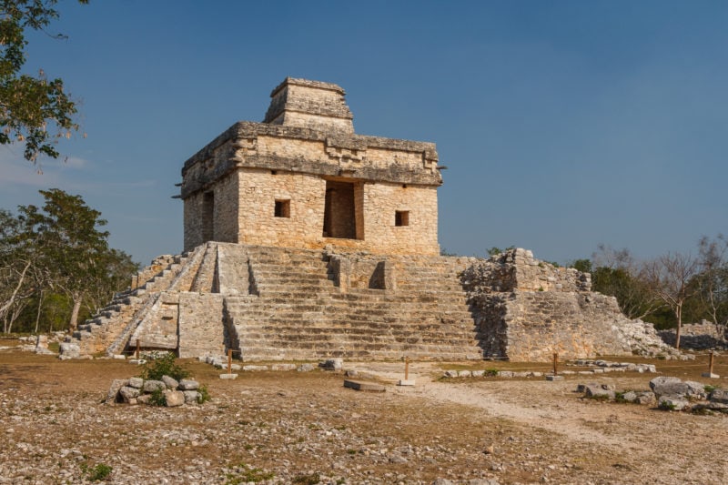 small ruined Mayan pyramid built from yellowish coloured stone with grey stone steps leading up to the temple. the land in front of the temple is arid and strewn with stones. 