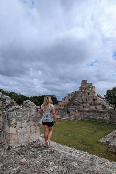 emily standing at the top of a grey stone mayan pyramid wearing blue denim shorts and a white top printed with palm trees with her blonde hair down. she is looking out across a grassy field towards a stone mayan pyramid on a grey day with a cloudy sky above at Edzna Archeological Site in Mexico