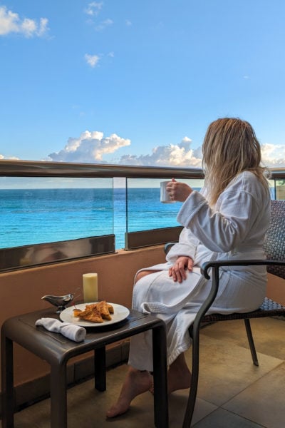 emily wearing a white bathrobe with her long blonde haid loose and messy. she is sitting on a hotel balcony next to a table laid with french toast and juice. she is holding a coffee and looking away from the camera towards the cyan coloured sea with blue sky above. 