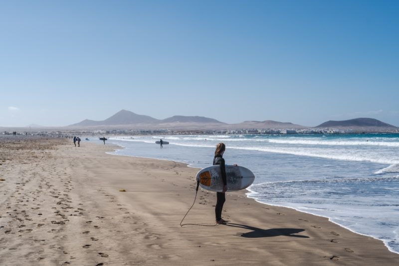 woman standing on a sandy beach holding a surfboard and looking at the sea with brown mountains in the background - best areas in lanzarote