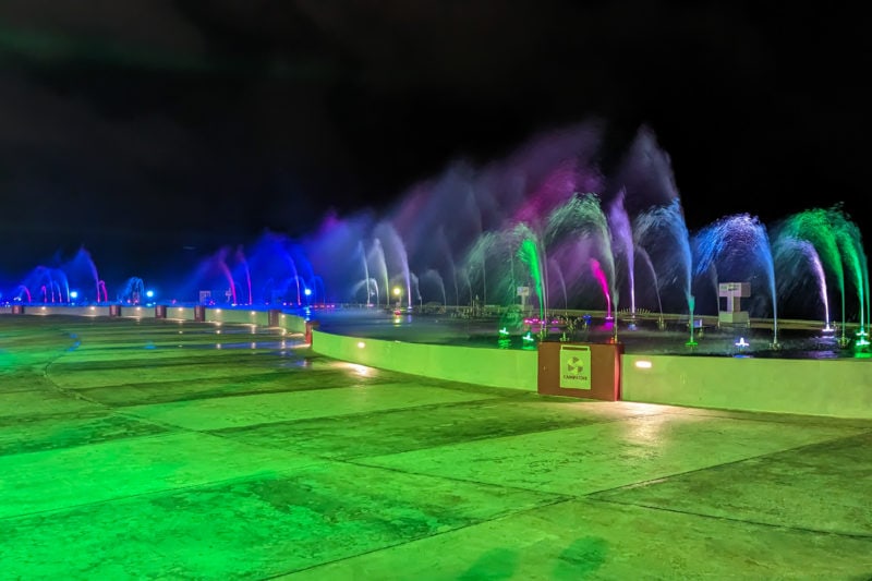 wide section of a paved esplanade at night with a long line of fountain jets all lit in different colours for the campeche fountain show