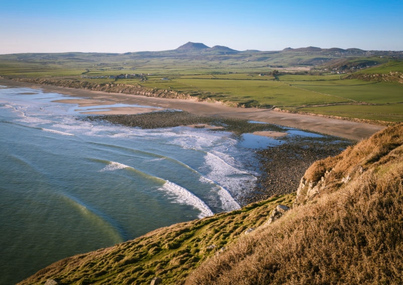 looking down at Hell's Mouth Beach, a small bay with a sandy beach and blue sea lined with white capped waves, with a green peninsula behind and mountains in the far distance, with a small patch of grassy headland visible in the foreground and blue sky overhead. best beaches on the llyn peninsula.