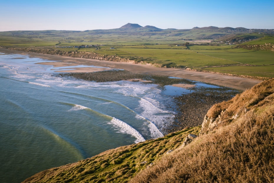 looking down at Hell's Mouth Beach, a small bay with a sandy beach and blue sea lined with white capped waves, with a green peninsula behind and mountains in the far distance, with a small patch of grassy headland visible in the foreground and blue sky overhead. best beaches on the llyn peninsula.