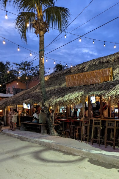 a bar with open front and wooden bar stools lined up along the edge of the road with a grass rood and a sign saying the Hot Corner at night with fairy lights overhead and a palm tree above in holbox mexico