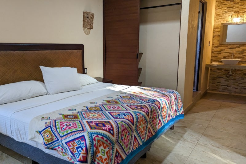 interior of a hotel room at Suites Villas el Jardín in Holbox with a large kingsized bed with white sheets and a mulicoloured throw with geometric design on the bottom of the bed. the room has grey tiled floors and white walls and there is an open wardrobe on the other side of the bed.