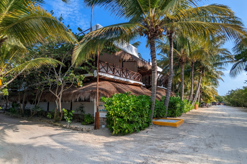 exterior of a two storey hotel building with whitewashed walls and a balcony all the way around the top floor with wooden railings. the hotel is mostly hidden behind palm trees and there is clear blue sky behind.